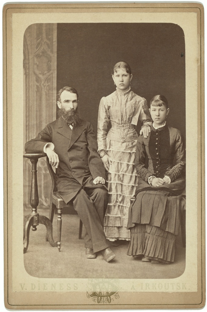 Archival sepia studio portrait with a seated man on the left, one daughter standing in the center and another seated on the right.